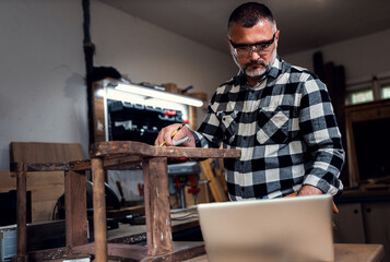 Carpenter standing in his workshop restoring old piece of furniture marking measurements on wood...