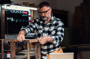 Carpenter standing in his workshop restoring old piece of furniture marking measurements on wood...
