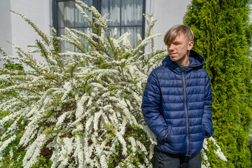 A young male wearing a blue puffer jacket stands by a flowering shrub in spring