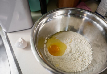 top view of a metal mixing bowl filled with flour and an egg on the surface with the shell on the kitchen counter