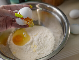 woman's hand cracking an egg over a metal mixing bowl filled with flour and another egg on the surface