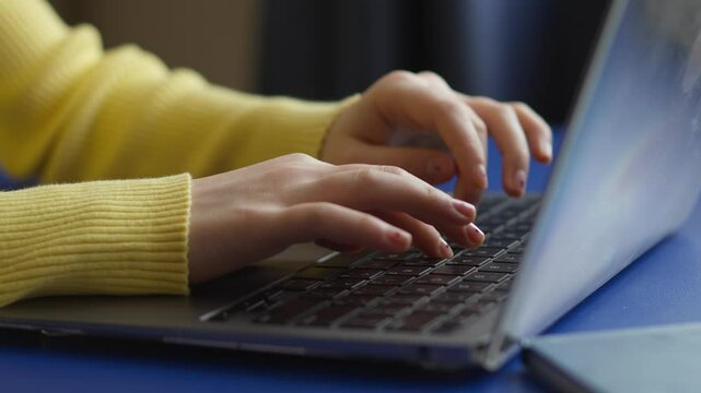 Close-up of hands typing on a laptop keyboard. A person wearing a yellow sweater works on a laptop with a bright blue surface. The image conveys a sense of productivity or communication.