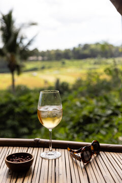 Cocktail with ice in a wine glass served with a dish of peanuts next to tortoise shell sunglasses with view of rice paddies in an open-air structure in Bali, Indonesia. 