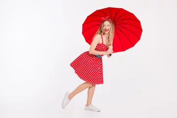 Fotobehang Cheerful young woman in a retro red polka dot dress holding a bright umbrella while posing gracefully against a white background. © deagreez