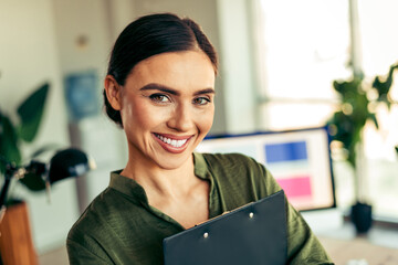 Confident young businesswoman smiling while holding documents in a modern workspace setting with...