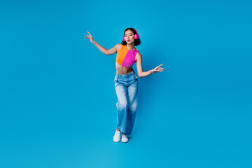 Young stylish woman dancing to music on blue backdrop wearing casual jeans and chic top with trendy...