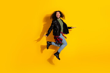 Young man with long curly hair jumps against a vibrant yellow background showcasing energy and fashionable style.