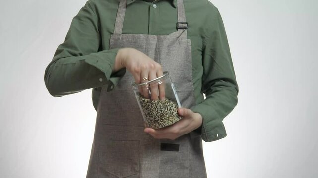 A man in a gardener's or povera apron holds a jar of seeds or spices, touches them and pours them into the jar.