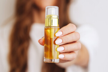 Woman showing hair serum. Hand holding glass bottle. Natural beauty care product. White background and blurred face. Focus on yellow oil. Clean skincare routine. Elegant and healthy lifestyle.