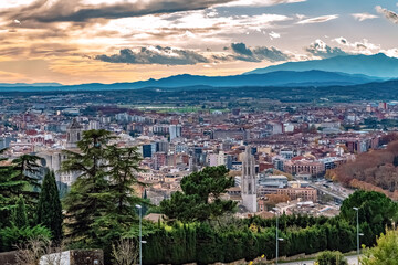 Fototapeta premium Panoramic view of Girona at sunset with church towers, historic and modern architecture, and the Pyrenees mountains in the background. The cityscape glows under a dramatic evening sky, Spain