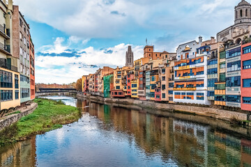 Girona cityscape in Spain featuring the Onyar River, colorful houses, and the Cathedral of Girona. Overcast sky with patches of blue and the reflections of buildings and sky in the water