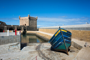 Traditional blue fishing boat on the pier near the historic Skala du Port tower in the harbor of Essaouira, Morocco. Sunny day with blue sky and calm water