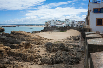 View from the ancient medina fortification in Essaouira, Morocco, capturing stone walls and whitewashed buildings beneath a cloudy sky. The Atlantic's rocky shoreline at low tide
