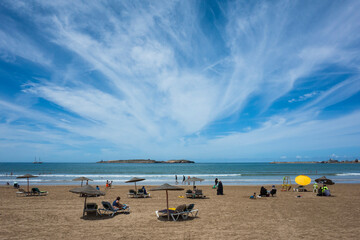 Sandy beach in Essaouira, Morocco, with sun loungers, umbrellas, people relaxing, and dramatic...