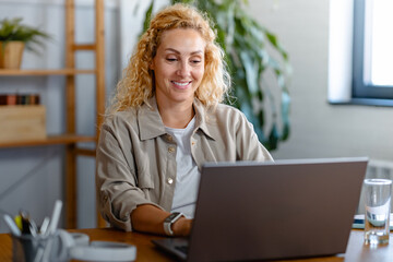 Smiling woman working on a laptop at a wooden desk in a cozy home office, enjoying a productive day in a relaxed and modern workspace.