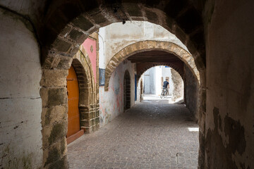 Obraz premium Stone archway in the medina of Essaouira, Morocco. A man rides a bicycle through the cobbled alley lined with historic walls and traditional architecture