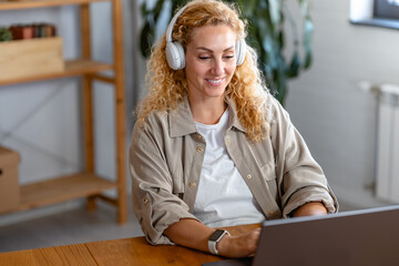 Smiling woman wearing headphones works on her laptop in a cozy home office setting, staying focused and engaged during an online meeting or remote work session.