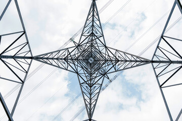 View of steel power tower from below against a cloudy sky, illustrating symmetrical structure pattern and lines  of energy transmission infrastructure