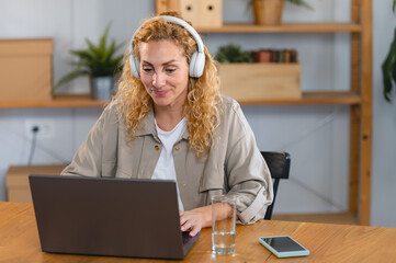 Focused young woman wearing headphones, working on laptop at home office desk with smartphone and glass of water, surrounded by plants.