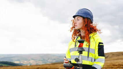 Portrait of confident female land surveyor with long hair in bright safety gear using surveying...