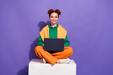Stylish young woman with red hair using a laptop seated on a modern block against a vibrant purple...