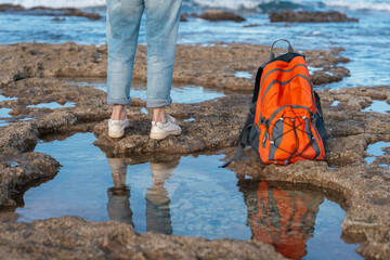 Backpacker explores rocky shoreline with vibrant orange backpack during daylight hours near the water