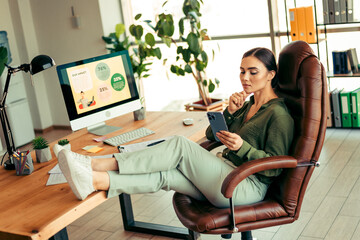 Confident Businesswoman Relaxing in a Modern Office Space with Phone and Computer, Representing...