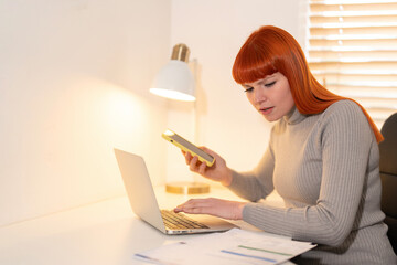 Young woman with red hair focuses on laptop while using smartphone in a well-lit home office during...