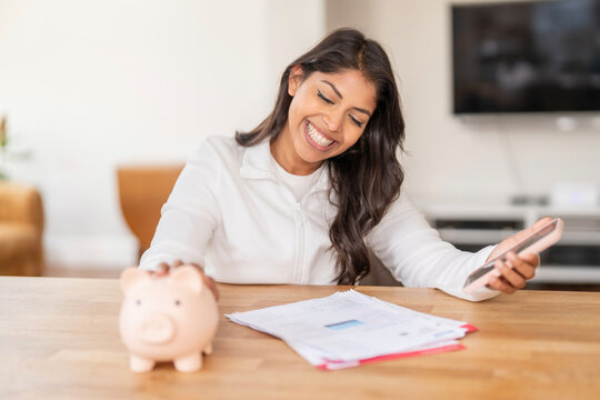 Young woman managing finances at home with piggy bank and smartphone while reviewing documents on a wooden table