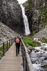Female hiker walks over a wooden boardwalk in a deep gorge, approaching Njupeskär Waterfall in Fulufjället National Park, surrounded by gray rocky cliffs and lush green in Dalarna Sweden