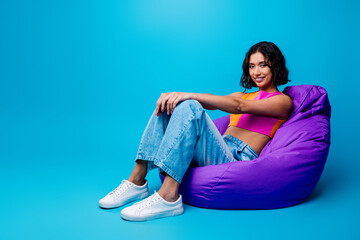 Joyful young woman sitting on a purple bean bag chair against a vibrant blue background showcasing...