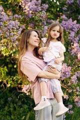 Fototapeta premium Mother holding her little daughter who makes a funny face while playing with her mom’s hair. Natural and candid family moment in a blooming spring garden with lilac flowers.
