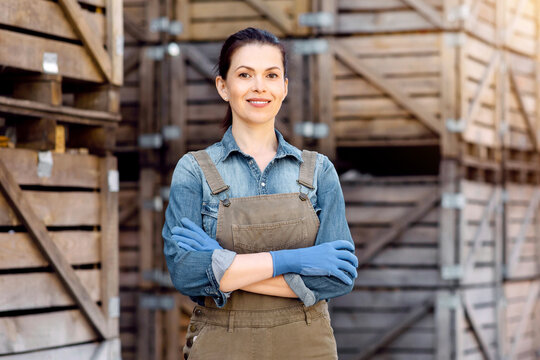 Owner of business, manager of storage and worker. Smiling confident millennial woman in uniform with crossed arms, on many wooden boxes background, shelves with goods in warehouse, free space