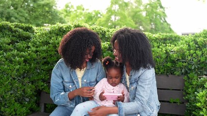 Two twin sisters enjoying time outdoors with their niece, sharing a smartphone on a park bench, creating a heartwarming family moment - Powered by Adobe