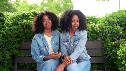Two twin sisters wearing denim jackets are holding hands while sitting together on a park bench near a green hedge - Powered by Adobe