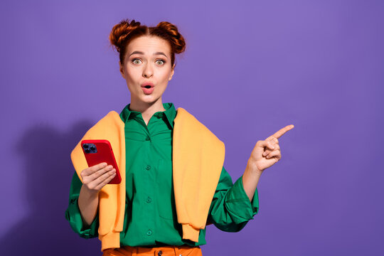 Cheerful red haired woman holding a smartphone while gesturing amazedly, standing against a vibrant violet backdrop