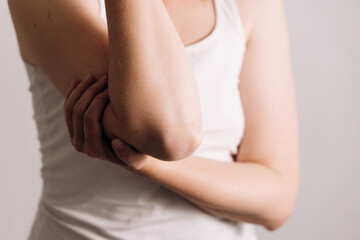 Close-up of a person with fair skin wearing a white tank top, gently holding their elbow with a thoughtful expression, conveying a sense of contemplation and introspection in a neutral background