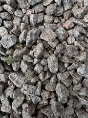 Close-up of rough gray granite gravel stones scattered on the ground, showing natural texture and irregular shapes with a hint of green plant growth.