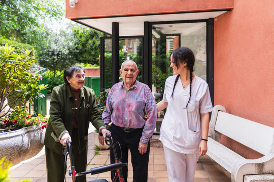 Nurse assisting elderly couple using walker in outdoor nursing home garden, providing support and care for seniors