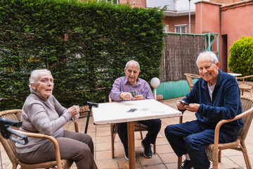 Three senior friends enjoying a card game outdoors in the garden of a nursing home, promoting social interaction and active aging