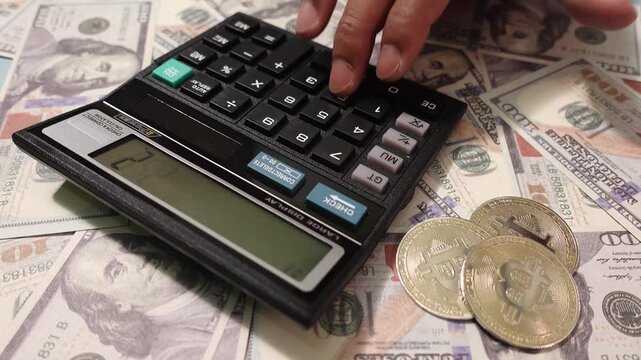 A hand operates a calculator surrounded by scattered US dollar bills and Bitcoin. Calculator and bitcoins in US dollars representing finances and cryptocurrencies