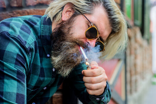 Bearded man in sunglasses smoking cigarette. Handsome bearded man lights cigarette. Closeup portrait of man with beard and mustache smoking on street. Cigarette smoke. Stylish hipster with cigarette.