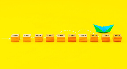 Wooden blocks with years and a blue paper boat on a yellow background representing future planning