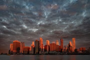 Fototapeta premium The Manhattan skyline rises imposingly beneath a dramatic sky, where clouds and the golden light of dawn intertwine in a scene of striking contrasts.
