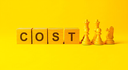 Cost spelled out with blocks and chess pieces on a yellow background in a studio setting
