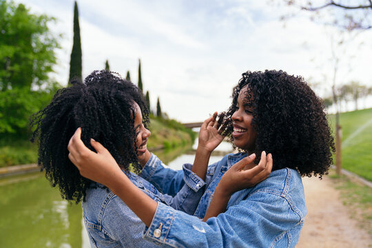 African two happy twin sisters are embracing and touching their curly afro hair, enjoying a sunny day together in a park - Powered by Adobe