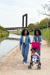 Twin sisters enjoying a walk in the park, pushing their niece in a stroller along a canal, creating...
