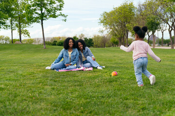 Two mothers sitting on the grass, enjoying the sight of their daughter joyfully running toward a vibrant, colorful ball