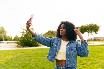 Young black woman adjusting her hair while taking a selfie with smartphone in a green park during a sunny day