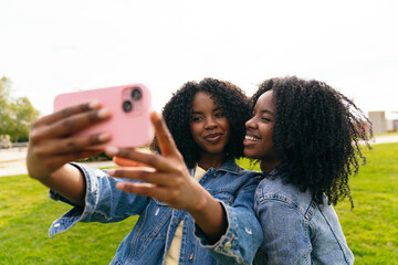 African two joyful twin sisters capturing a cheerful selfie with a smartphone in a vibrant park, both wearing trendy denim jackets